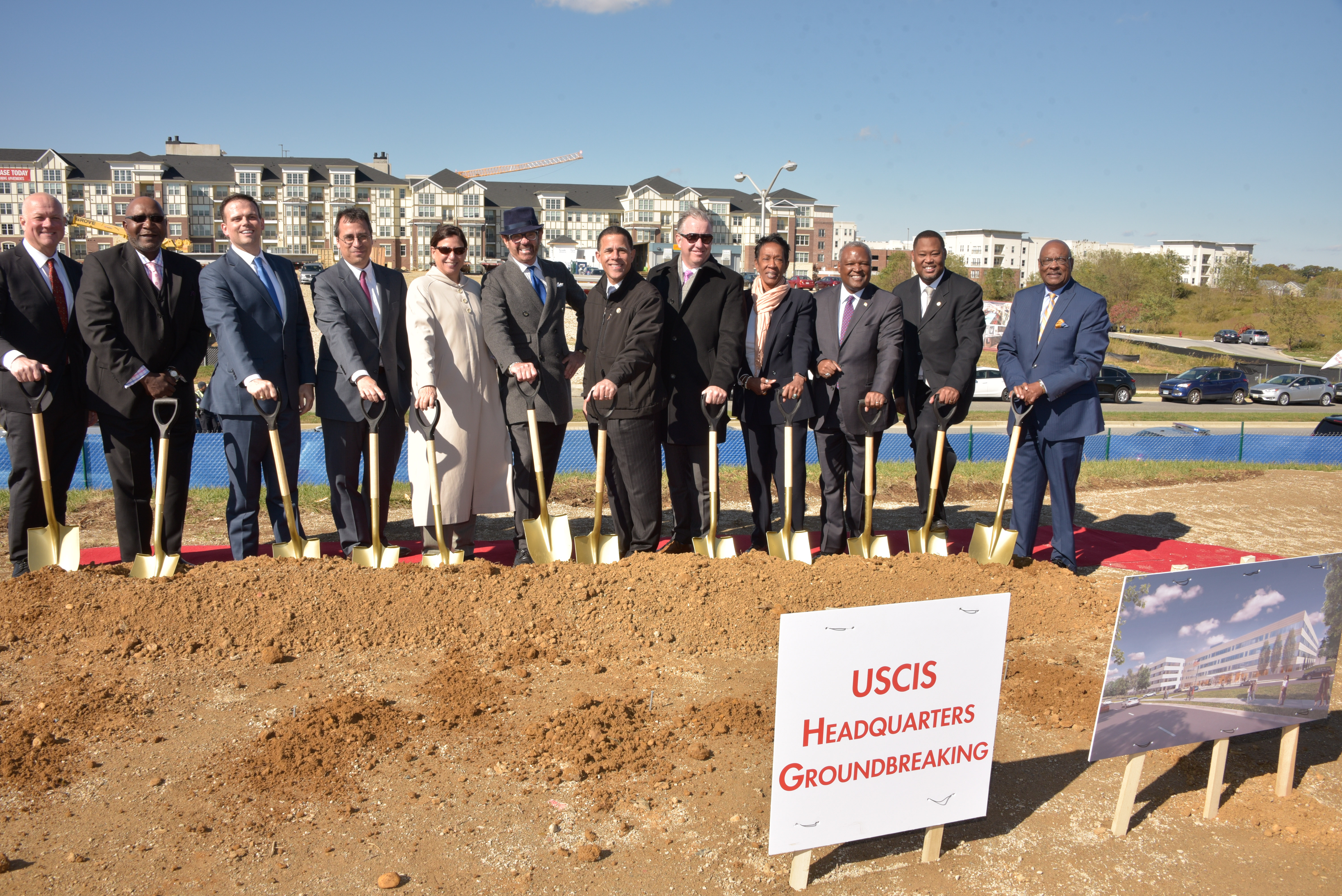 Ceremonia de inicio de construcción de la sede de USCIS en Camp Springs, Maryland, con un cartel de USCIS en primer plano.